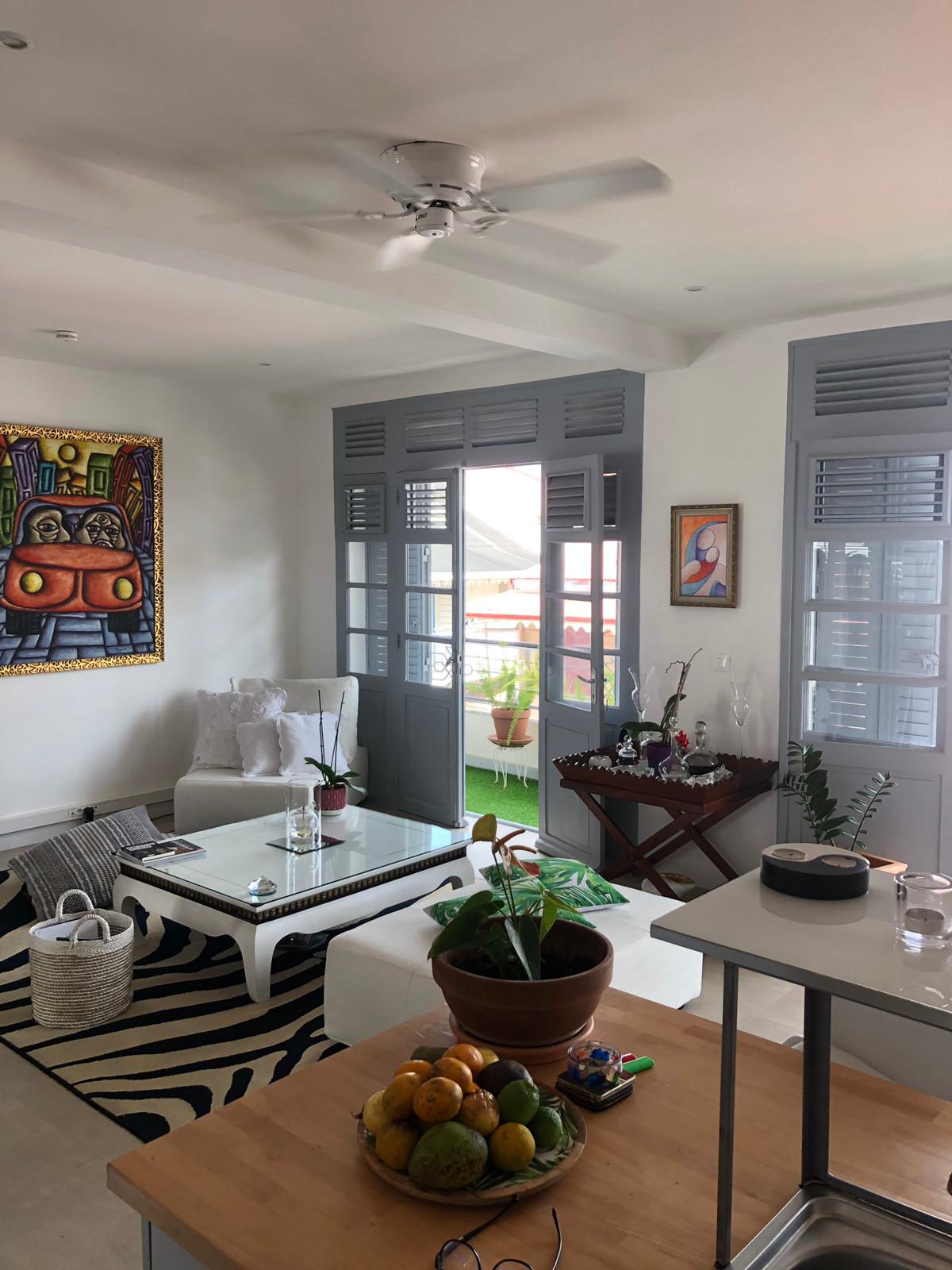 Int&eacute;rieur d'un appartement r&eacute;nov&eacute; : vue du salon depuis la cuisine ouverte et des fen&ecirc;tres ext&eacute;rieures &agrave; clayettes, caract&eacute;ristiques de la Guadeloupe