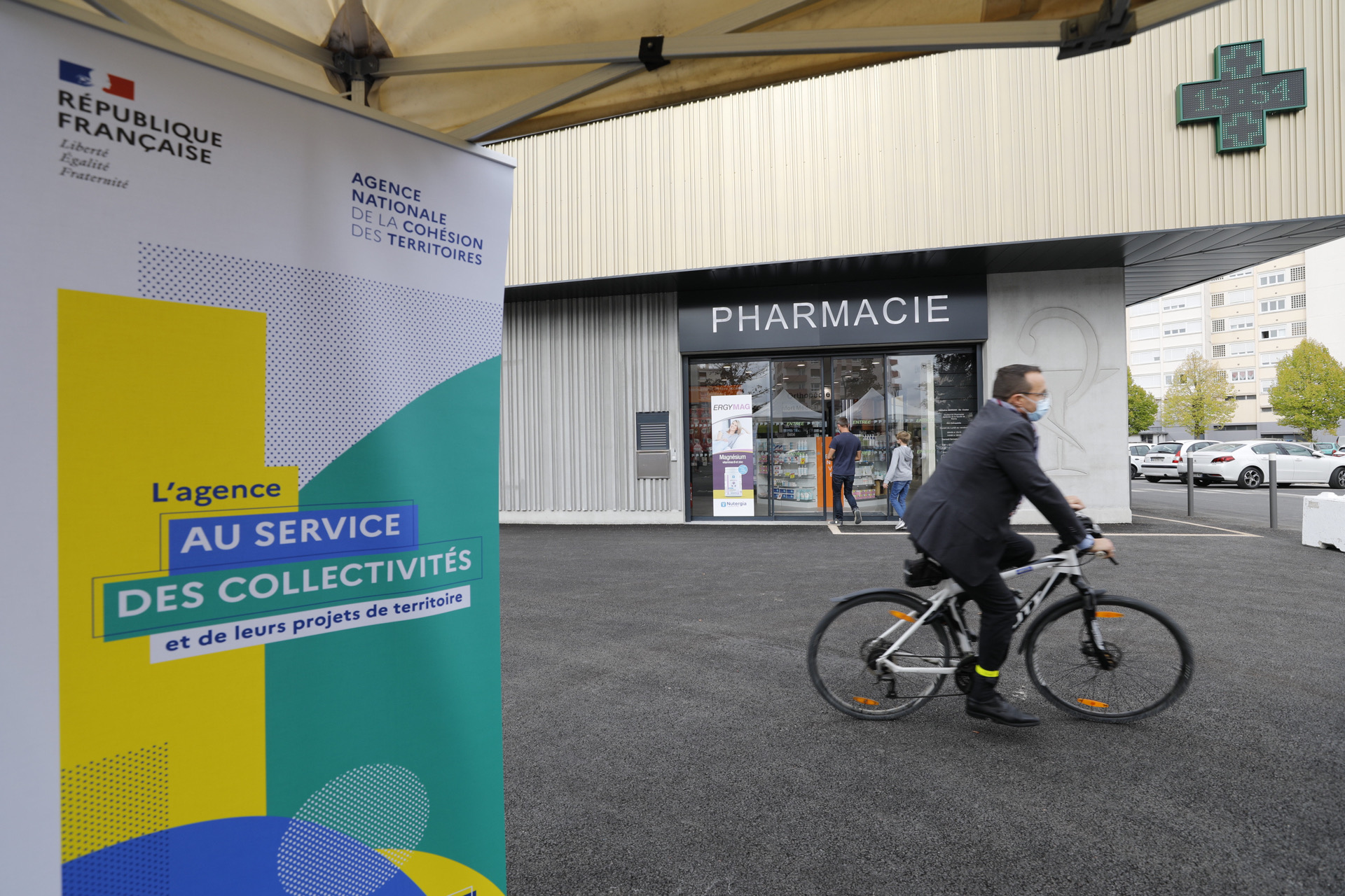Kak&eacute;mono de l'ANCT devant le centre comercial, avec un monsieur &agrave; v&eacute;lo qui passe devant la nouvelle pharmacie