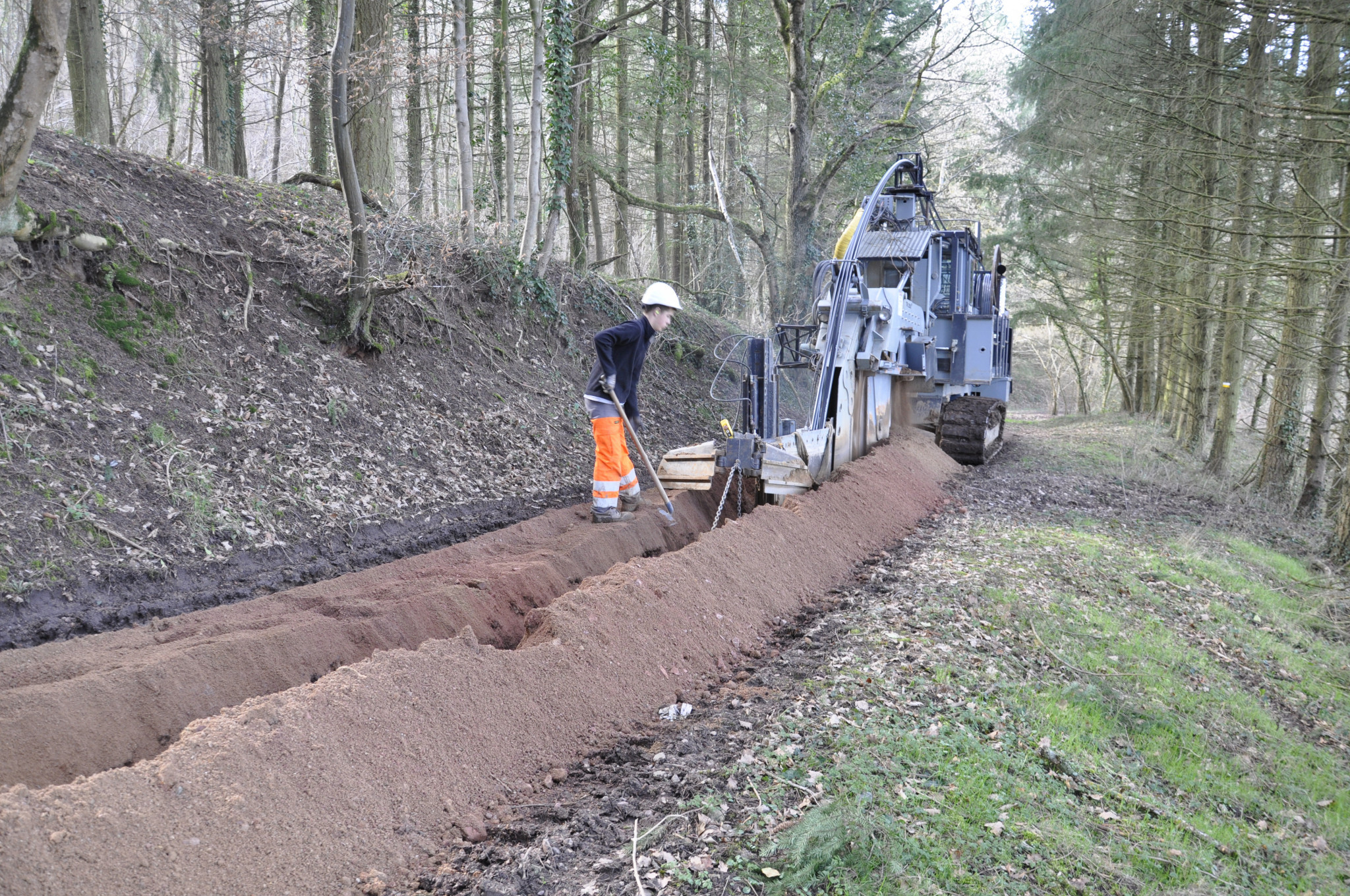 Installation de la fibre avec une machine qui creuse un foss&eacute; et un ouvrier qui d&eacute;plpoie la ligne, dans un sous-bois