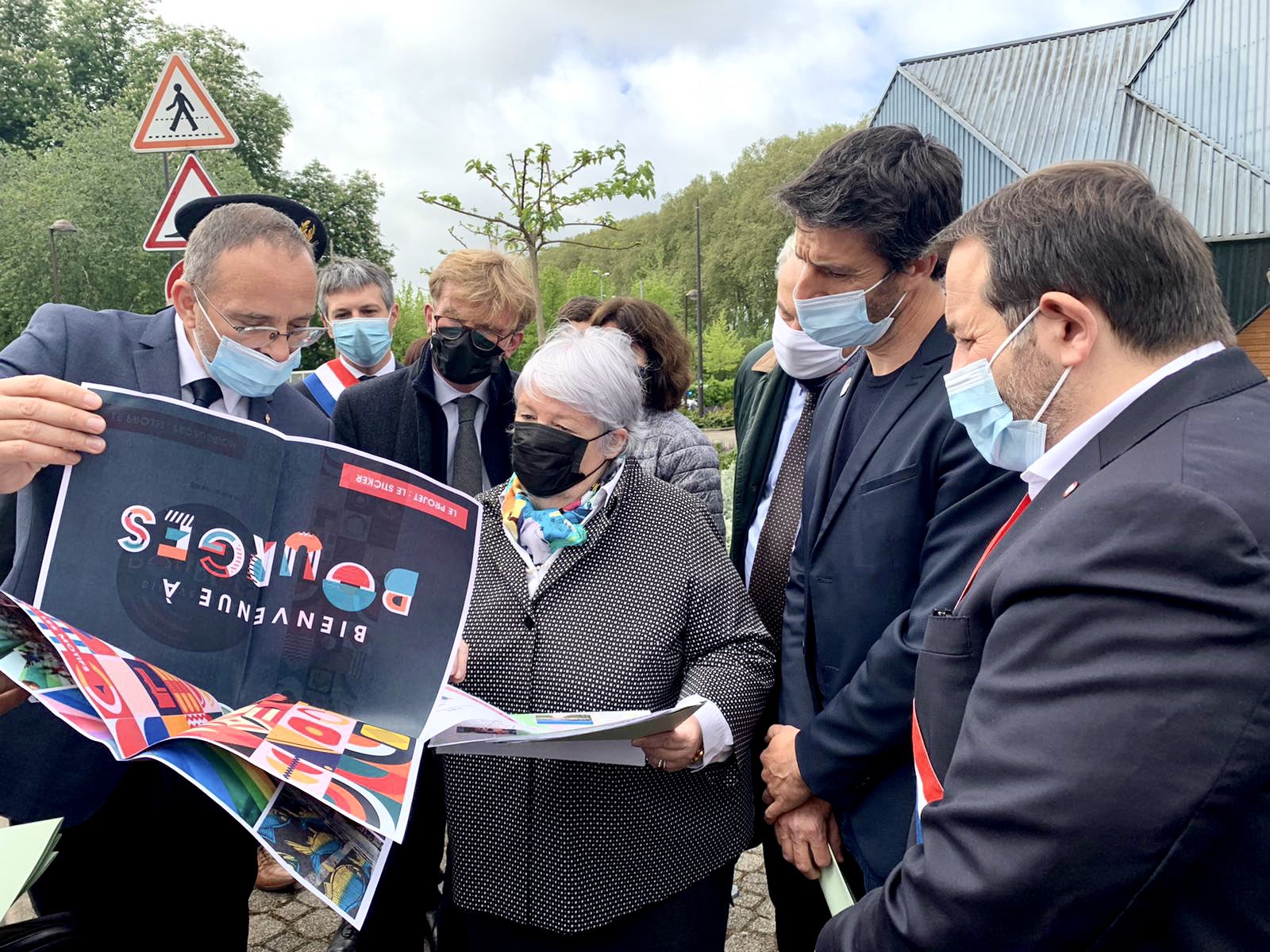 En ext&eacute;rieur, groupe de personnes, en costume et avec le masque, qui entoure la ministre Jacqueline Gourault pour lui montrer une affiche du Forum sur le desing actif. 