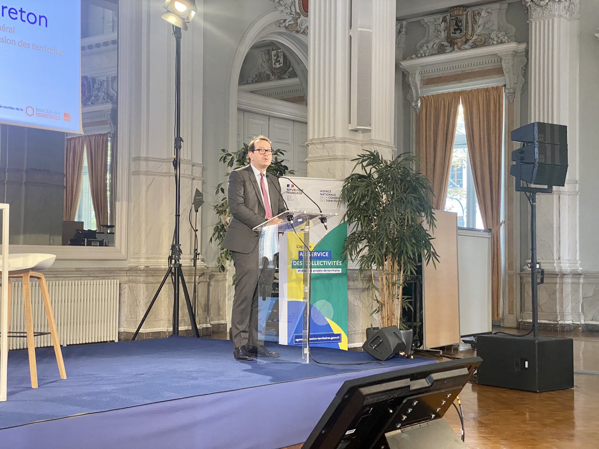 Yves Le Breton en costule gris est au pupitre, sur une estrade au tapis mauve, pour faire son discours