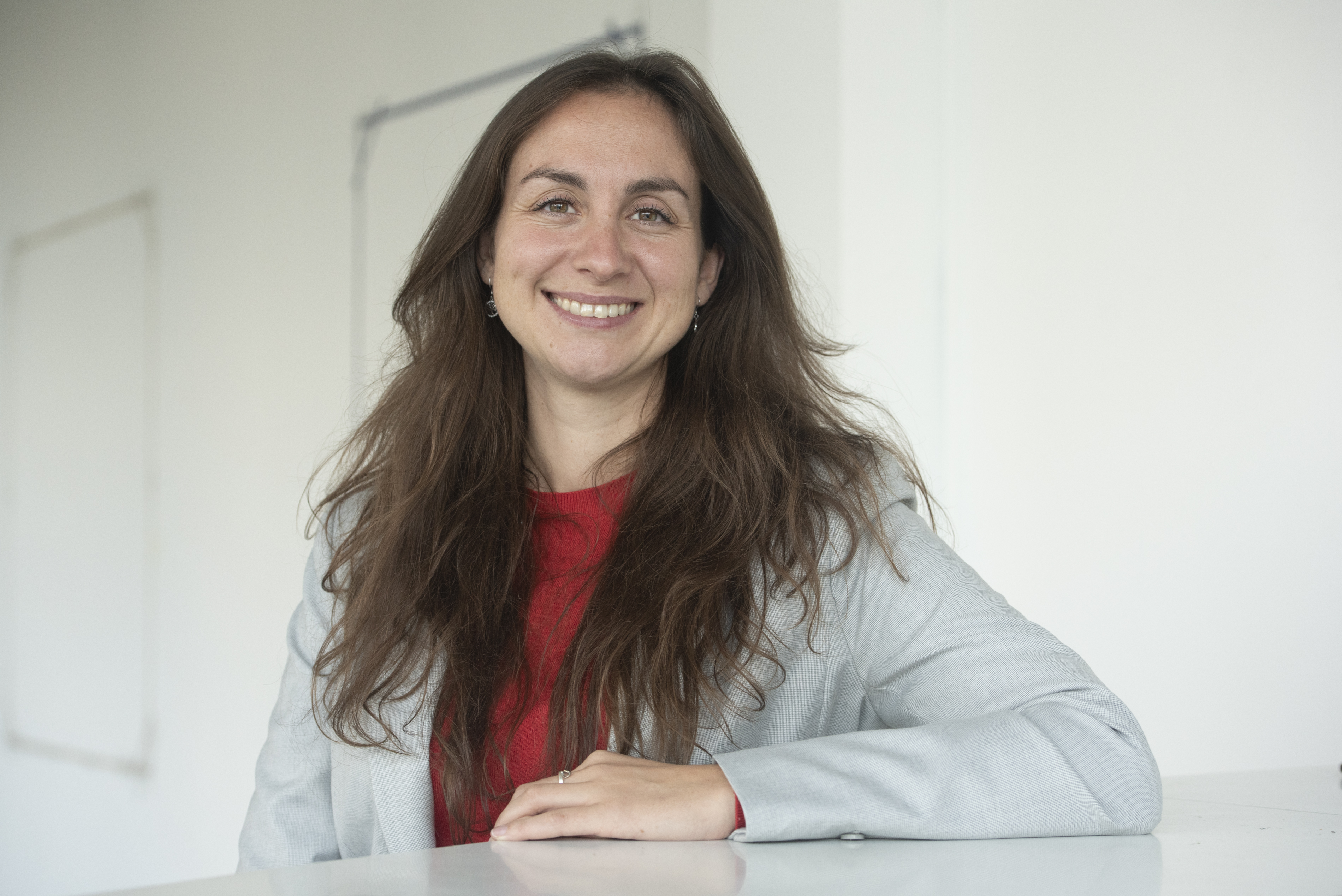 Portrait d'Emilie Schmitt, souriante, cheveux chataings longs, blouson gris clair ouvert sur un pull rouge 
