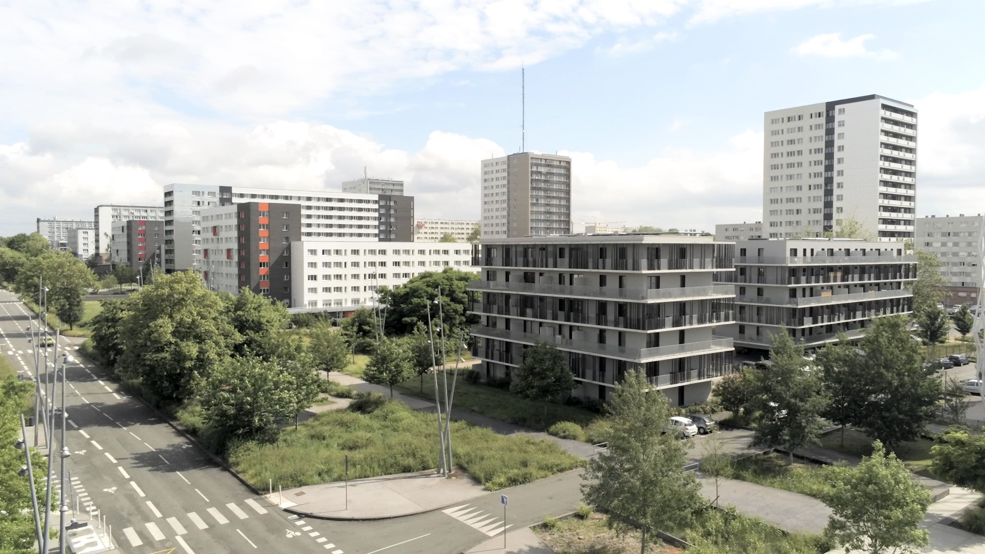 Vue sur une grande avenue arborée, des immeubles d'habitations modernes et, au second plan, de grandes tours blanches.