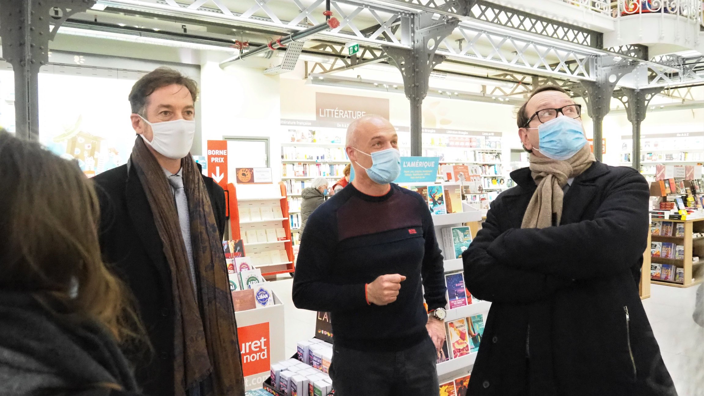 Dans une librairie avec des poutes métalliques, 3 hommes regardent l'architecture du lieu.