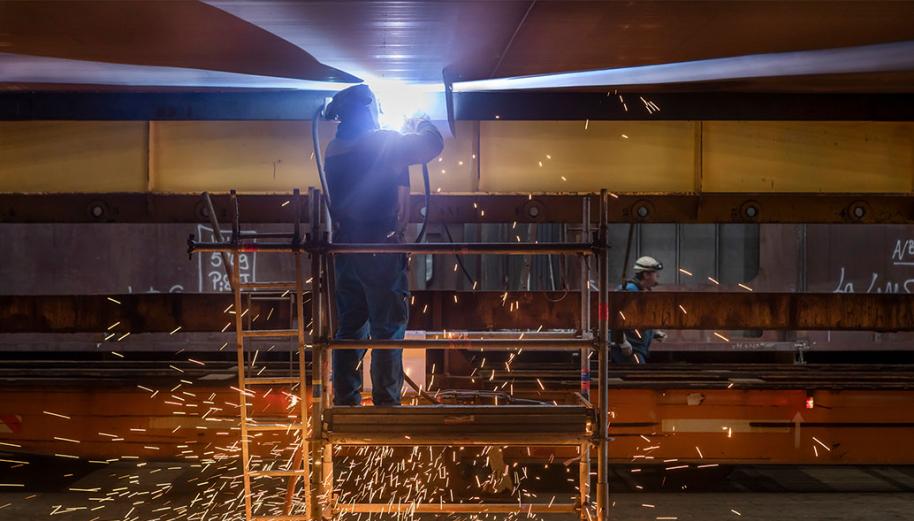 Sur un échaffaudage, un homme en bleu de travail et avec un casque de protection est en train de souder un tuyau. On le voit de dos, entouré de gerbes d'étincelles.