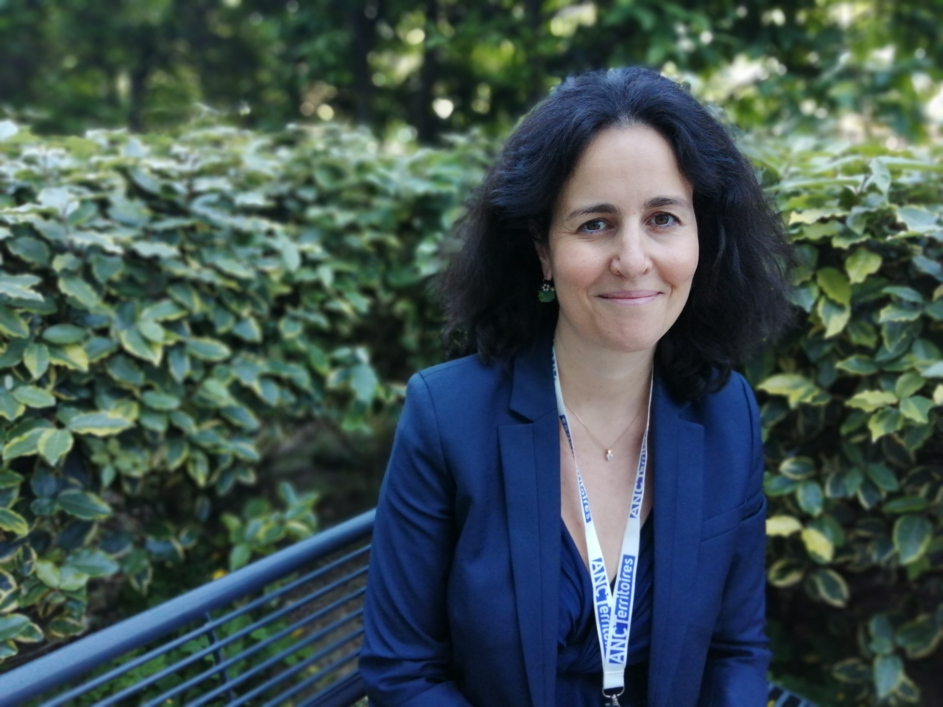 Portrait en extérieur, sur un banc enturé de haies ; femme brune, cheveux mi-longs en tailleur bleu foncé