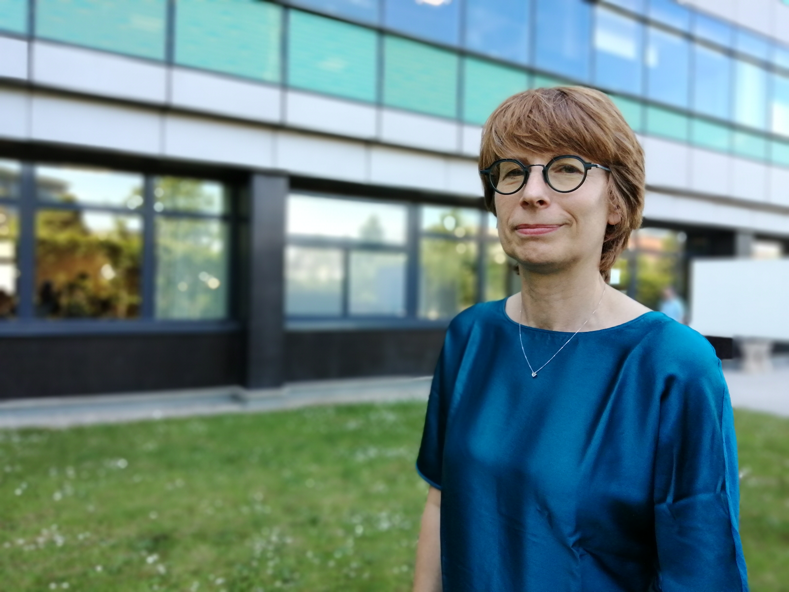Portrait extérieur devant un bâtiment en verre ; lunette, cheveux courts et chemisier bleu