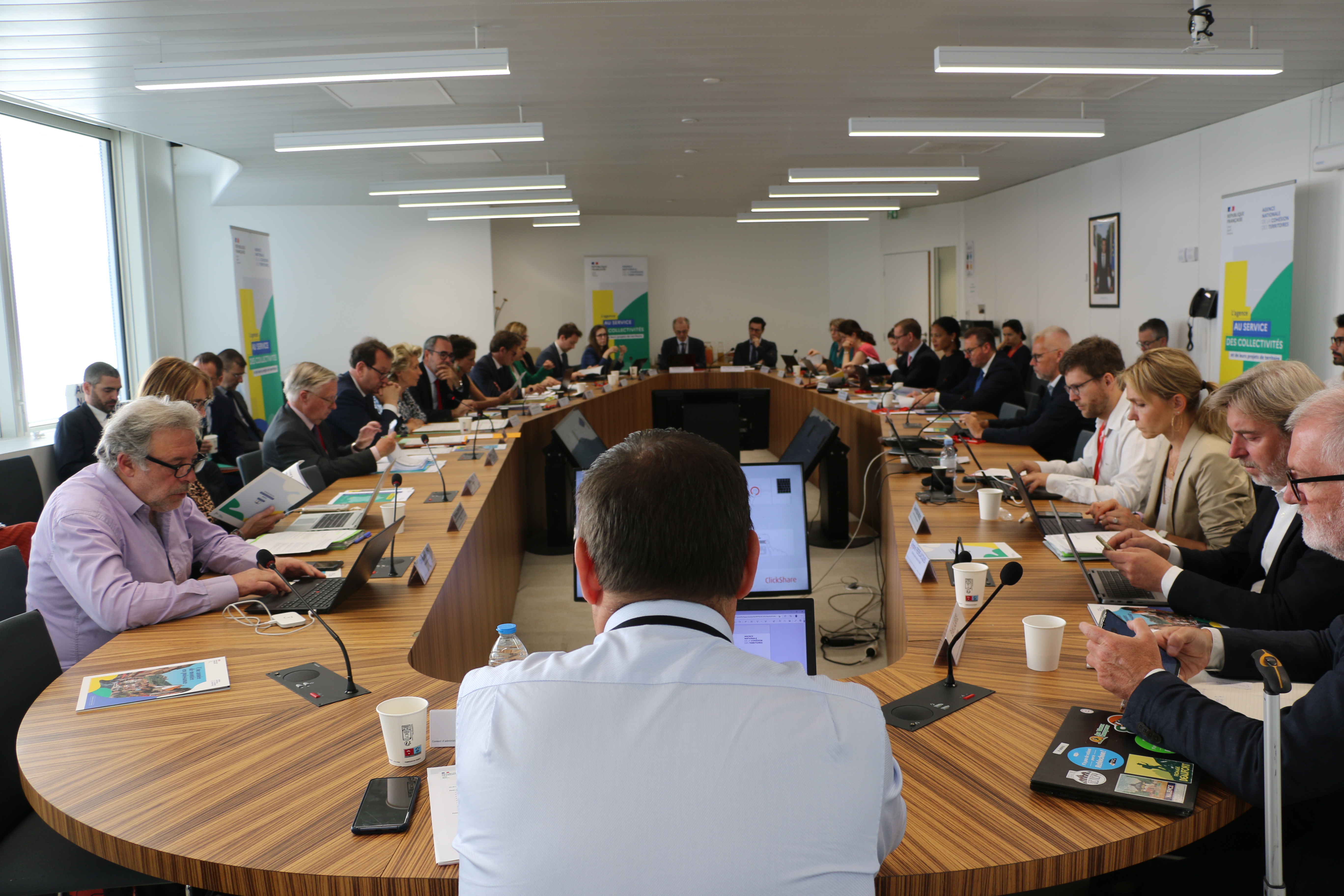 VUe de l'ensemble des membres et invit&eacute;s du CA, autour de 2 grandes rang&eacute;es de tables, disposes en ovale. De dos, un homme en chemise blanche, au 1er plan.