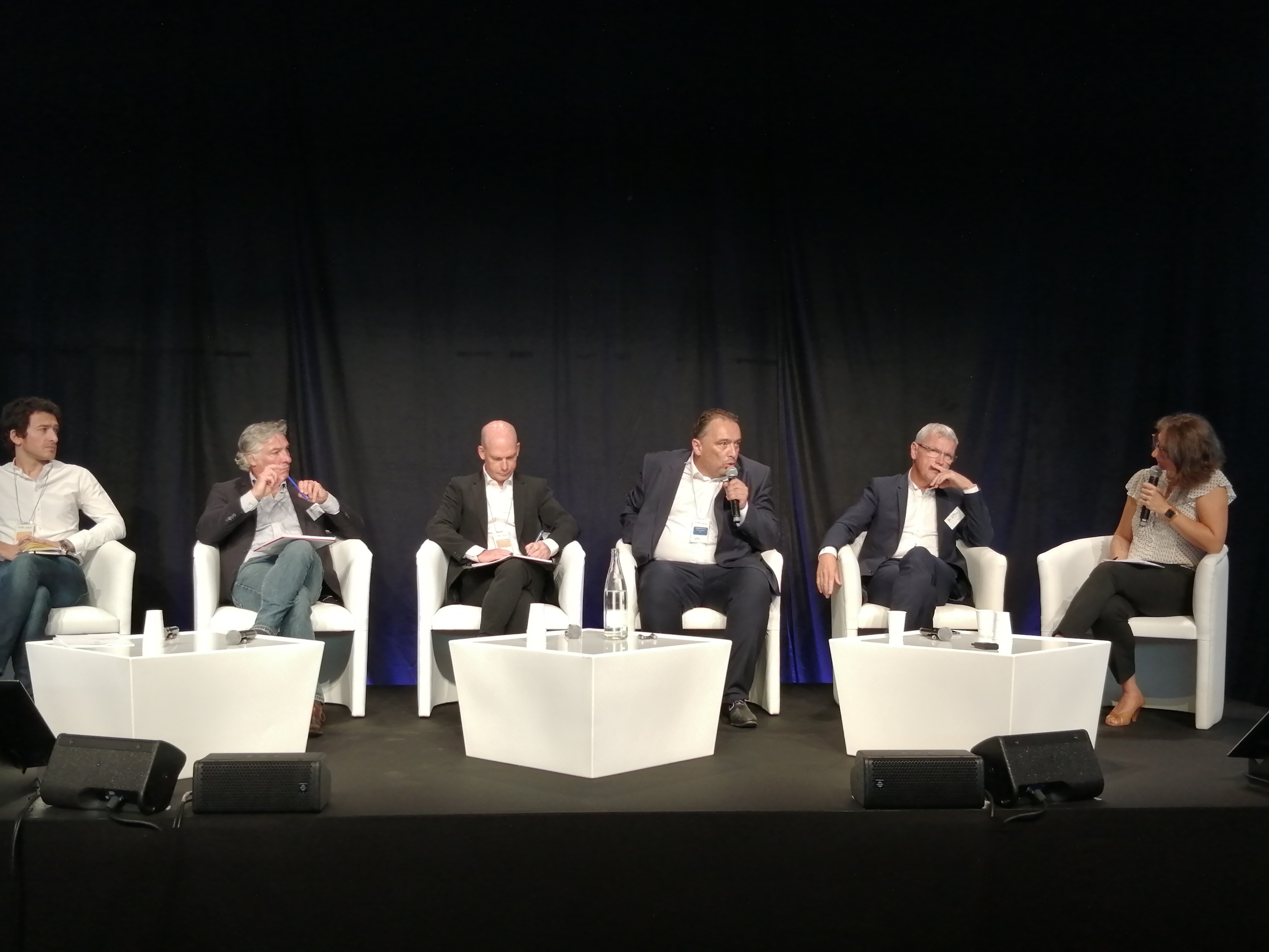 Vue de l'estrade où sont assis plusieurs participants à un débat, dans des fauteuils blancs.