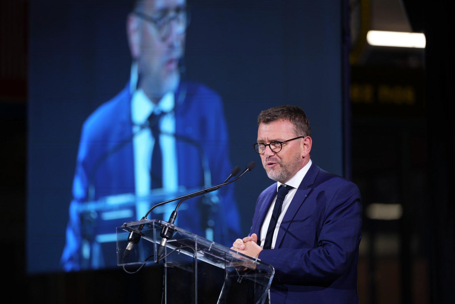 Olivier Klein, en train de parler devant un pupitre avec micro, en costume bleu foncé et avec un grand écran derrière lui