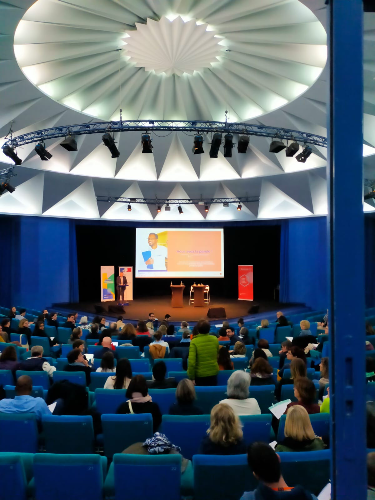 Vue de l'arrière d'un grand amphithéâtre avec des fauteuils bleus, face à la scène qui se trouve au fond. Au plafond, un grand luminaire blanc en forme de rond.