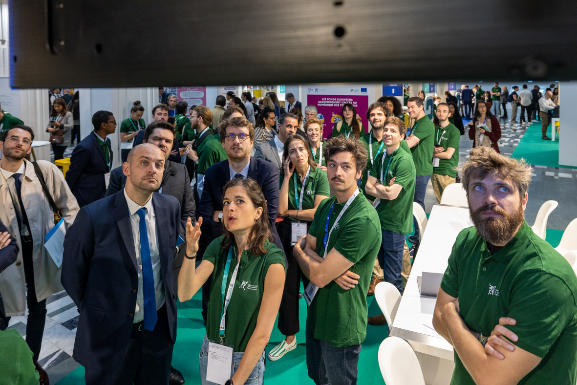Vue en plong&eacute;e sur des agents ANCT en t-shirt vert qui pr&eacute;sentent qq chose au ministre JL Barrot. 