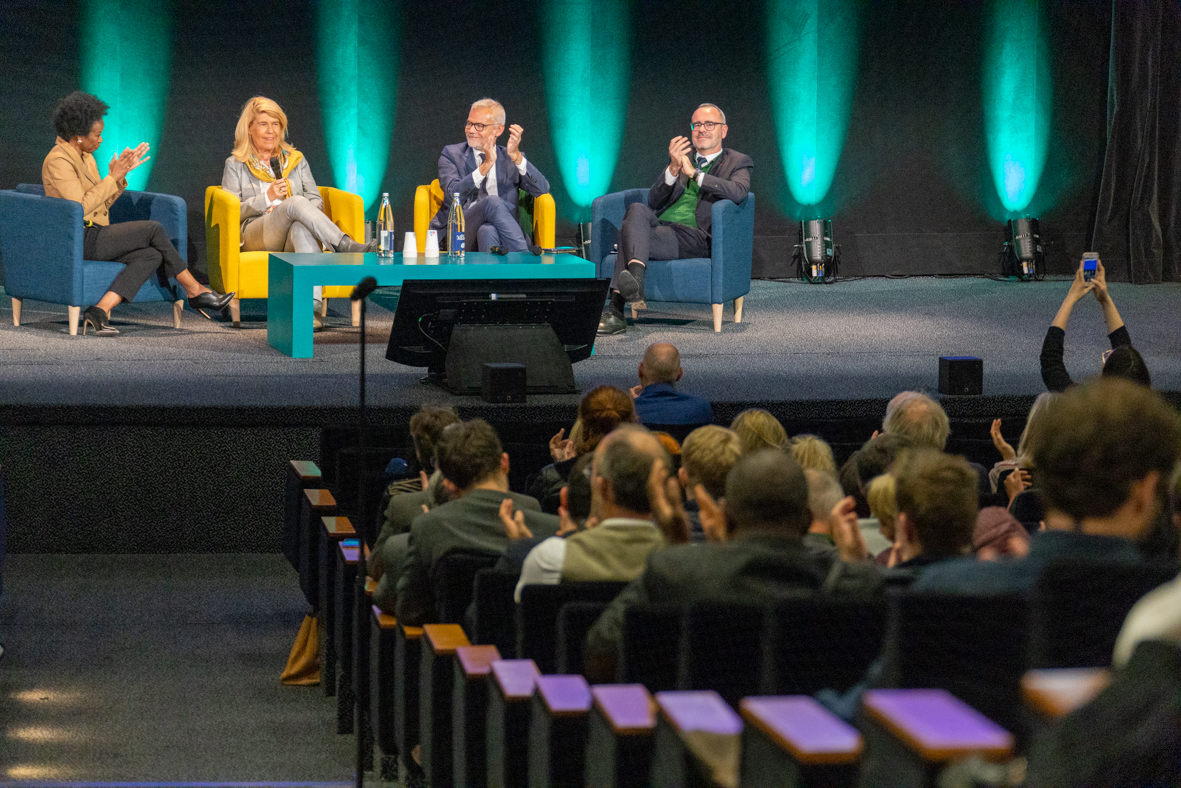 Vue d'un amphit&eacute;ha&acirc;tre, avec des personnes assises de dos et, sur sc&egrave;ne, en fond, quatre personnes de face.