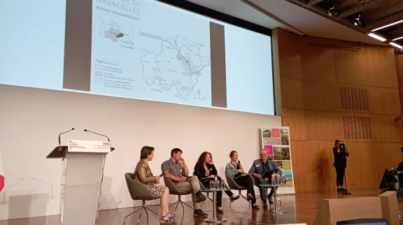 Sur une sc&egrave;ne des personnes  intervenantes sont align&eacute;es, avec un grand &eacute;cran au-dessus d'elles.